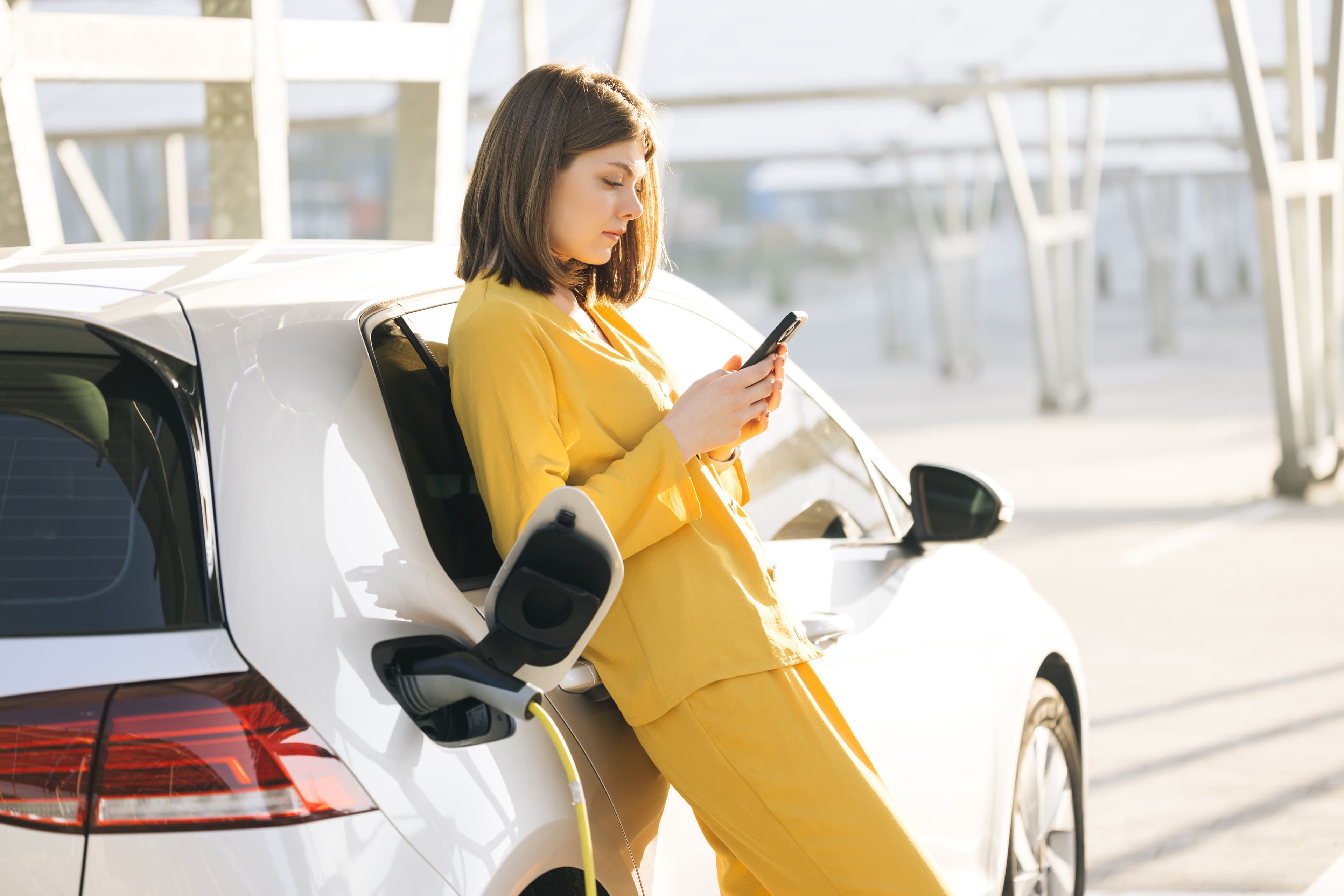 Frau lehnt im orangenen Hosenanzug am Elektroauto blick auf das Handy und wartet bis das Auto geladen ist.